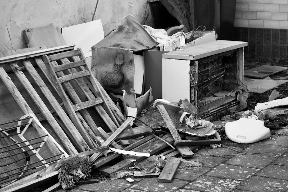 A collection of discarded household items and debris piled against an exterior wall in a cluttered outdoor space. The pile includes a weathered wooden garden ladder leaning partially against the wall,