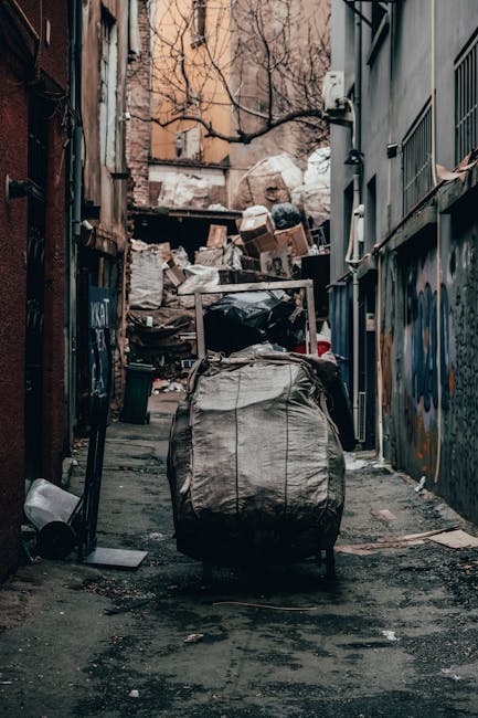 A collection of discarded household items and debris piled against an exterior wall in a cluttered outdoor space. The pile includes a weathered wooden garden ladder leaning partially against the wall,