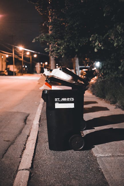 A black wheeled rubbish bin labeled 'ST. JOHN'S' is positioned on the edge of a sidewalk in a residential or urban area during nighttime. The bin contains multiple layers of waste, including cardboard